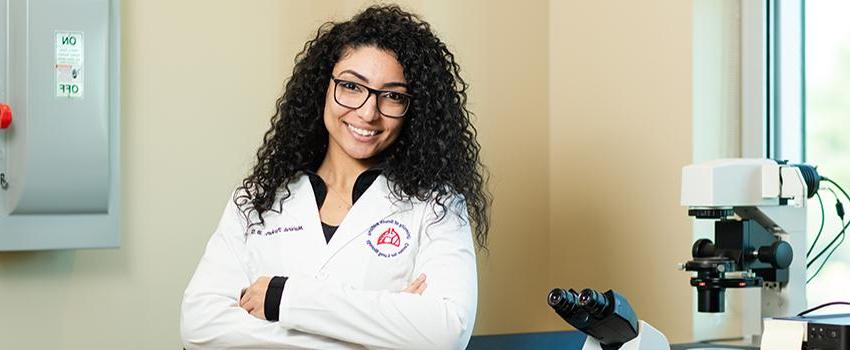 Female student in white lab coat with microscope behind her.